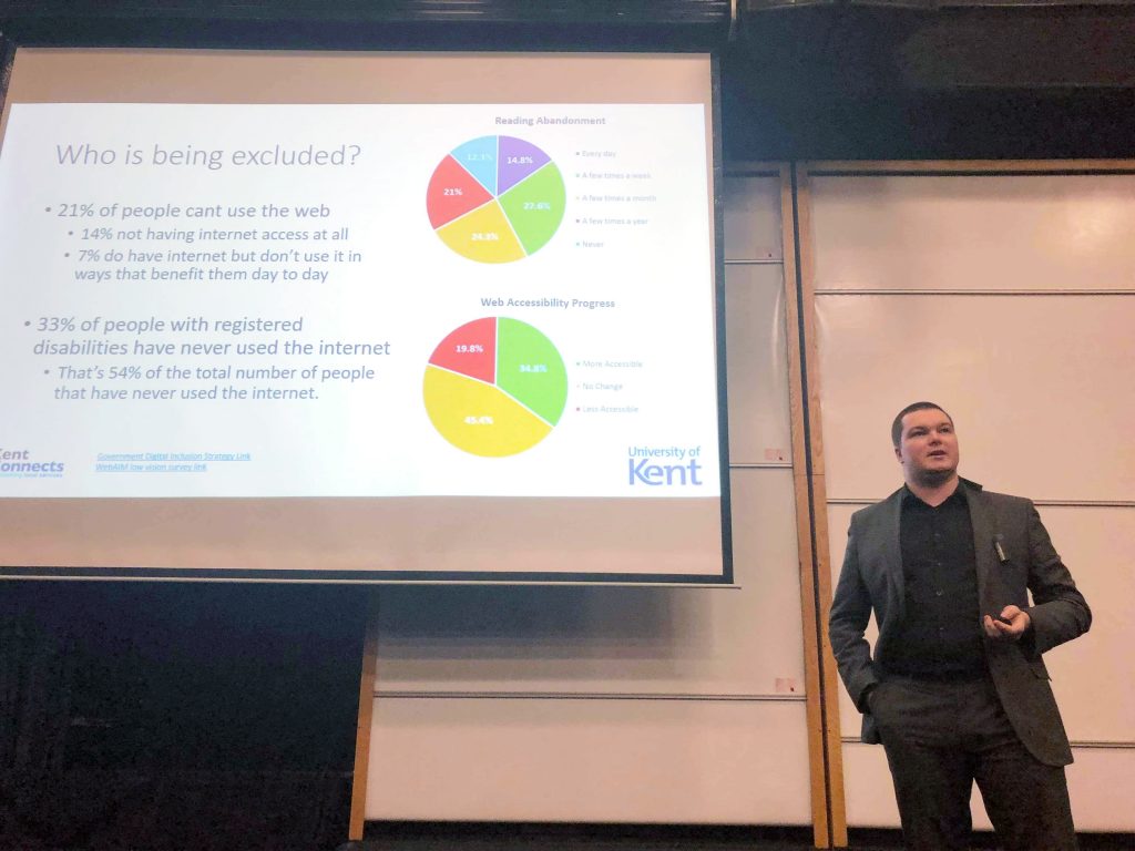 An image of George Rhodes, KCC Digital Accessibility Compliance Lead, giving a lecture at the University of Kent on Accessibility and its importance for students is the services they build. The picture shows George standing next to his slides, which are displaying figures on the levels of digital exclusion in the UK. The slides shown are available to download elsewhere on this page.